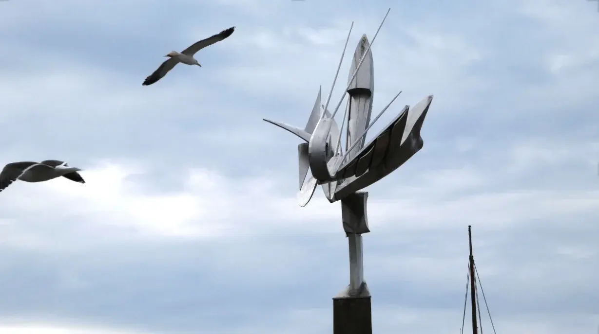 Stille bevegelse: blank metallskulptur som folder seg ut som vinger mot en skyet himmel, med to måker i flukt